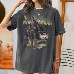 Op de afbeelding: Een antracietgrijs t-shirt met korte mouwen en een grafische print van The Mandalorian en Grogu. Het ontwerp bevat de tekst "STAR WARS THE MANDALORIAN" en "THIS IS THE WAY" in een verweerd lettertype met gedempte, aardse kleurtinten.