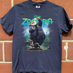 K&ouml;nnte beinhalten: Ein dunkelblaues Kurzarm-T-Shirt mit einem Grafikdruck der Figur Clawhauser aus dem Disney-Film Zoomania. Die Figur tr&auml;gt eine Polizeiuniform und ist vor dem leuchtend gr&uuml;nen Zoomania-Filmlogo abgebildet.