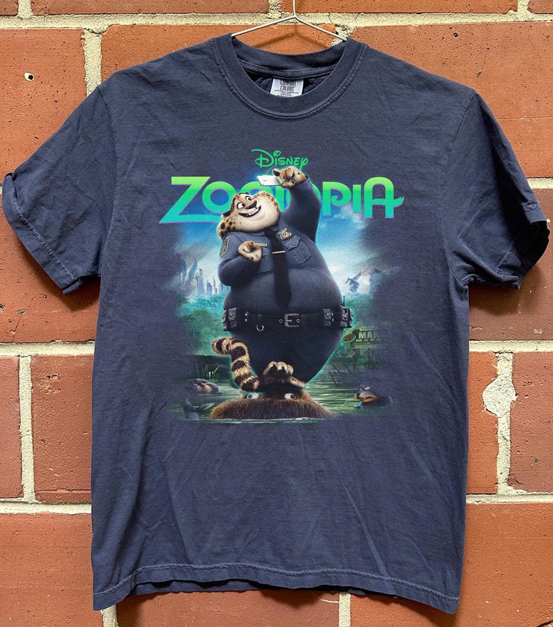 K&ouml;nnte beinhalten: Ein dunkelblaues Kurzarm-T-Shirt mit einem Grafikdruck der Figur Clawhauser aus dem Disney-Film Zoomania. Die Figur tr&auml;gt eine Polizeiuniform und ist vor dem leuchtend gr&uuml;nen Zoomania-Filmlogo abgebildet.