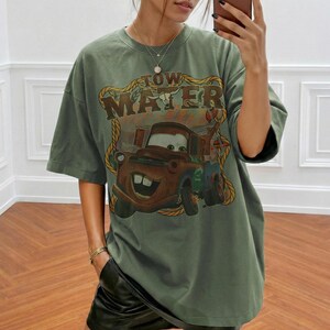 May include: Olive green t-shirt featuring a graphic of Tow Mater from the movie Cars. The design includes the text "Tow Mater" in a brown, distressed font. The shirt has short sleeves and a relaxed fit.