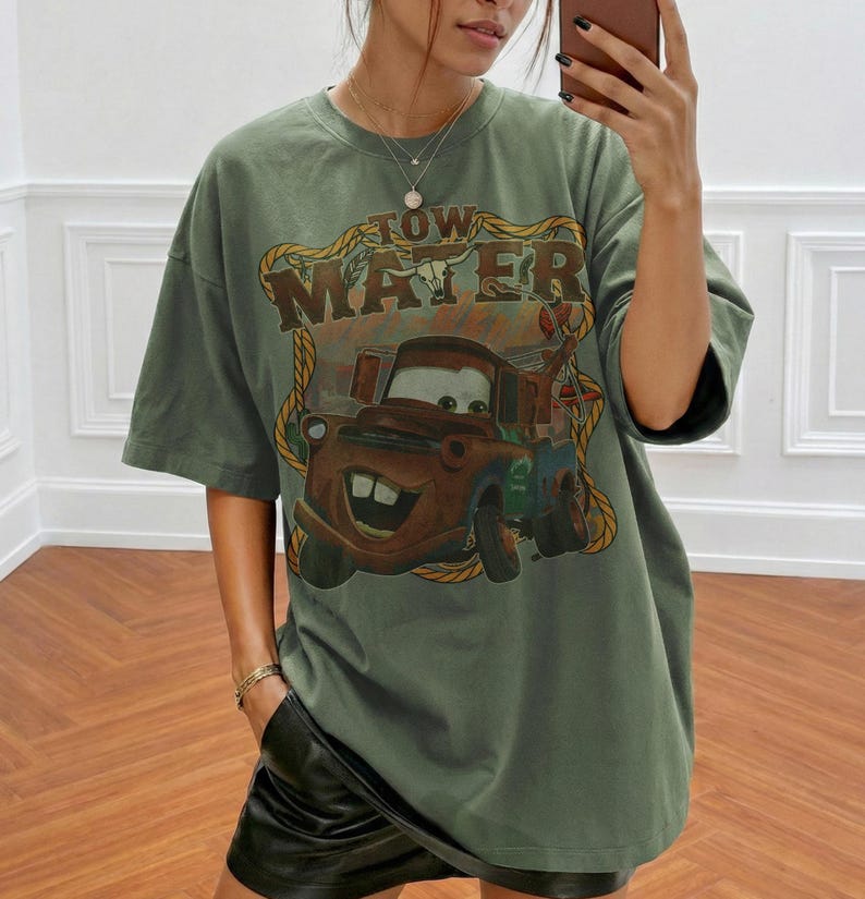 May include: Olive green t-shirt featuring a graphic of Tow Mater from the movie Cars. The design includes the text "Tow Mater" in a brown, distressed font. The shirt has short sleeves and a relaxed fit.