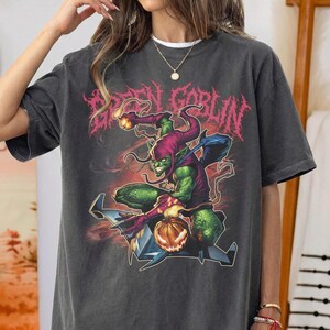 May include: Dark gray t-shirt featuring a Green Goblin graphic. The design includes the text "GREEN GOBLIN" in pink, a green goblin character, and a jack-o'-lantern. The shirt has short sleeves and a crew neck.