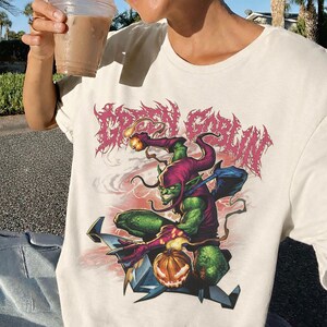 May include: Off-white t-shirt featuring a Green Goblin graphic. The design includes a green goblin character in a purple and red outfit, holding a pumpkin and a bomb. The words "GREEN GOBLIN" are printed above the character in a pink, stylized font.