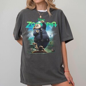 K&ouml;nnte beinhalten: Ein anthrazitfarbenes Kurzarm-T-Shirt mit einem Grafikdruck der Figur Clawhauser aus Zoomania in Polizeiuniform. Der Schriftzug Zoomania ist in leuchtendem Gr&uuml;n &uuml;ber der Figur auf einem blau-gr&uuml;nen Hintergrund aufgedruckt.