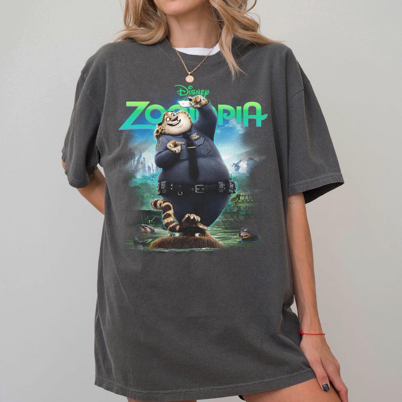 K&ouml;nnte beinhalten: Ein anthrazitfarbenes Kurzarm-T-Shirt mit einem Grafikdruck der Figur Clawhauser aus Zoomania in Polizeiuniform. Der Schriftzug Zoomania ist in leuchtendem Gr&uuml;n &uuml;ber der Figur auf einem blau-gr&uuml;nen Hintergrund aufgedruckt.