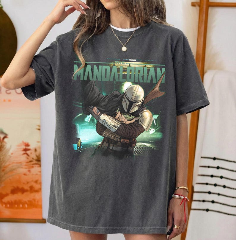 Puede incluir: Una camiseta de manga corta gris carb&oacute;n con un estampado gr&aacute;fico del personaje Mandalorian sosteniendo a una peque&ntilde;a criatura verde. El texto "MANDALORIAN" est&aacute; impreso en letras verdes grandes sobre el personaje. Cuello redondo.
