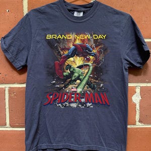 May include: A dark blue short-sleeve t-shirt featuring a graphic print of Spider-Man fighting a green creature. The text "BRAND NEW DAY" appears in yellow above the characters, and the red and blue "SPIDER-MAN" logo is printed at the bottom.