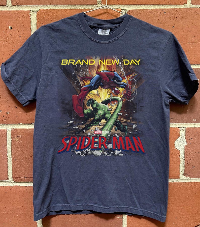 May include: A dark blue short-sleeve t-shirt featuring a graphic print of Spider-Man fighting a green creature. The text "BRAND NEW DAY" appears in yellow above the characters, and the red and blue "SPIDER-MAN" logo is printed at the bottom.