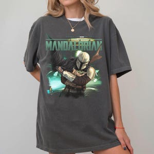 Puede incluir: Una camiseta de manga corta gris carb&oacute;n con un estampado gr&aacute;fico de un personaje con casco y armadura met&aacute;lica que sostiene a una peque&ntilde;a criatura verde. El texto "MANDALORIAN" est&aacute; impreso en letras may&uacute;sculas verdes sobre la ilustraci&oacute;n.