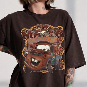 May include: Brown t-shirt featuring a graphic of Tow Mater from the movie Cars. The design includes the text "Tow Mater" in a Western-style font, with a cartoon image of the character. The shirt has a vintage, distressed look.