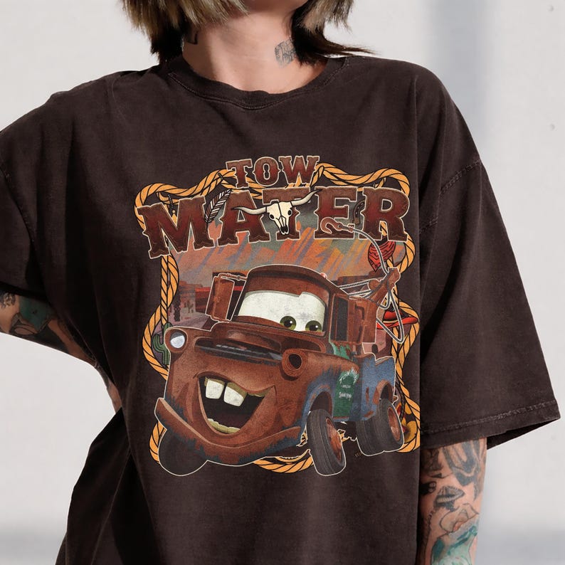May include: Brown t-shirt featuring a graphic of Tow Mater from the movie Cars. The design includes the text "Tow Mater" in a Western-style font, with a cartoon image of the character. The shirt has a vintage, distressed look.
