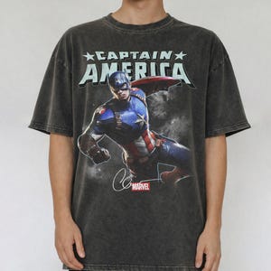 Puede incluir: Camiseta gris oscuro con Captain America. El gr&aacute;fico incluye el texto "CAPTAIN AMERICA" sobre una imagen del superh&eacute;roe en acci&oacute;n. El logotipo de Marvel y la firma tambi&eacute;n son visibles. La camiseta tiene un aspecto vintage.