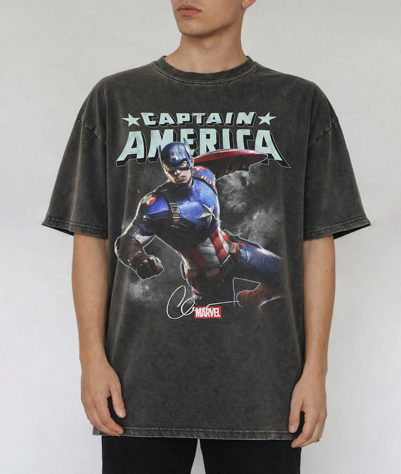 Puede incluir: Camiseta gris oscuro con Captain America. El gr&aacute;fico incluye el texto "CAPTAIN AMERICA" sobre una imagen del superh&eacute;roe en acci&oacute;n. El logotipo de Marvel y la firma tambi&eacute;n son visibles. La camiseta tiene un aspecto vintage.