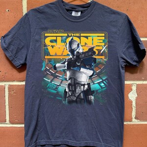 May include: A dark blue short-sleeve t-shirt featuring a graphic print of a white and blue clone trooper holding a blaster rifle. Above the character, the text reads STAR THE CLONE WARS in yellow and white letters against a rectangular frame.