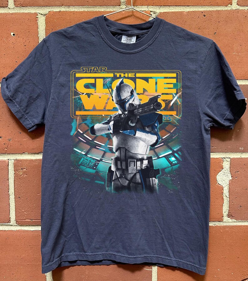 May include: A dark blue short-sleeve t-shirt featuring a graphic print of a white and blue clone trooper holding a blaster rifle. Above the character, the text reads STAR THE CLONE WARS in yellow and white letters against a rectangular frame.