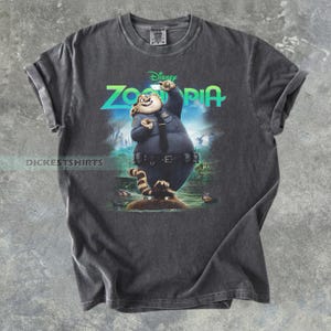 K&ouml;nnte beinhalten: Ein anthrazitfarbenes Kurzarm-T-Shirt mit einem Grafikdruck der Zootopia-Figur Officer Clawhauser. Die Figur tr&auml;gt eine blaue Polizeiuniform und h&auml;lt ein Telefon. Das Wort Zootopia ist in leuchtendem Gr&uuml;n dar&uuml;ber aufgedruckt.