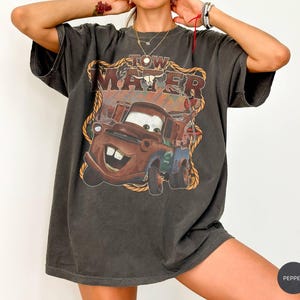 May include: Dark gray t-shirt featuring a graphic of Tow Mater from the movie Cars. The design includes the text "Tow Mater" in a western-style font, with a brown and orange color scheme. The shirt has a vintage, worn look.