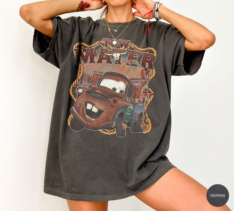 May include: Dark gray t-shirt featuring a graphic of Tow Mater from the movie Cars. The design includes the text "Tow Mater" in a western-style font, with a brown and orange color scheme. The shirt has a vintage, worn look.