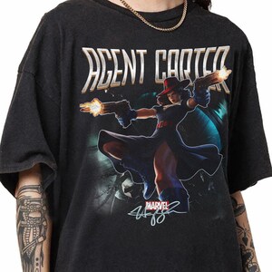Pu&ograve; includere: T-shirt nera con grafica di Agent Carter della Marvel, che tiene due pistole con fiamme. Il testo "AGENT CARTER" &egrave; sopra il personaggio. Il logo Marvel e la firma sono in basso.