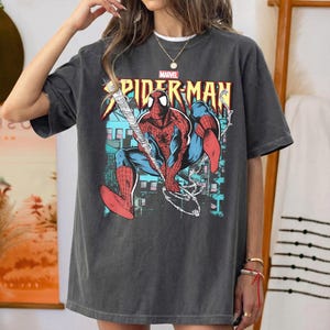 May include: A charcoal gray short-sleeve t-shirt featuring a graphic print of the Marvel character Spider-Man in a crouching pose. The design includes the text Spider-Man in yellow and red letters, with a web-slinging action scene in the background.
