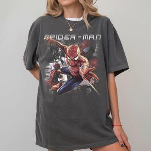 May include: A charcoal gray short-sleeve t-shirt featuring a graphic print of the character Spider-Man in a dynamic pose. The text SPIDER-MAN is printed in white above the character, with additional faded imagery of the character in the background.