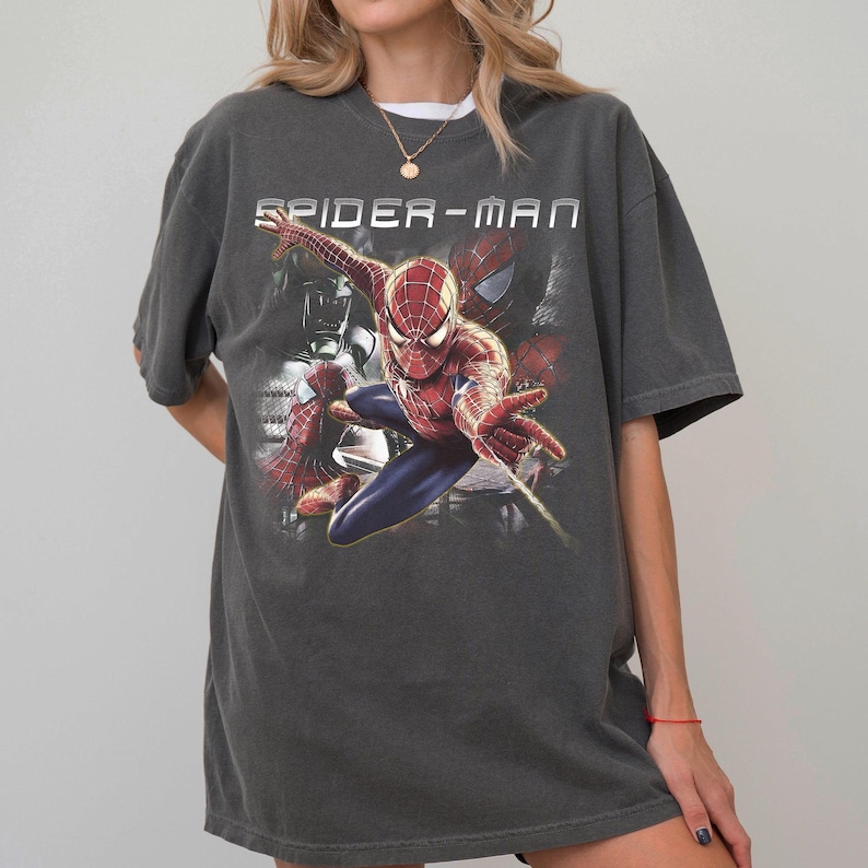 May include: A charcoal gray short-sleeve t-shirt featuring a graphic print of the character Spider-Man in a dynamic pose. The text SPIDER-MAN is printed in white above the character, with additional faded imagery of the character in the background.