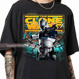 May include: A black short-sleeve t-shirt featuring a graphic print of a white and blue clone trooper holding a blaster. The design includes the yellow text STAR THE CLONE WARS and a background of a futuristic space station interior with teal lights.