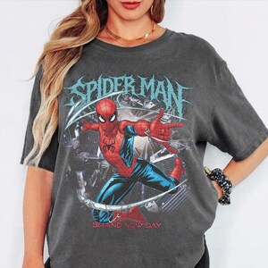 May include: Dark gray t-shirt featuring a graphic of Spider-Man in a dynamic pose, with the words "SPIDER MAN" in a stylized font. The phrase "BRAND NEW DAY" is printed below the image.