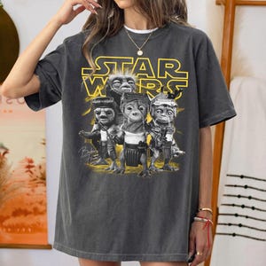 Op de afbeelding: Een antracietgrijs t-shirt met korte mouwen voorzien van een geel Star Wars-logo boven een afbeelding van drie kleine buitenaardse wezens. De wezens zijn afgebeeld in een monochrome stijl met gedetailleerde texturen en de tekst Babu Frik staat eronder.
