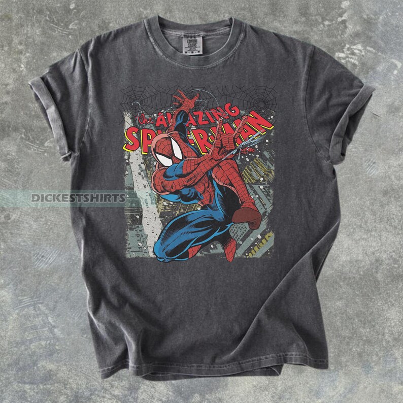 Puede incluir: Una camiseta gris carb&oacute;n de manga corta con un estampado gr&aacute;fico del personaje de c&oacute;mic Spider-Man en una pose din&aacute;mica. El dise&ntilde;o incluye el texto rojo y amarillo The Amazing Spider-Man, un fondo de ciudad y patrones de telara&ntilde;a.