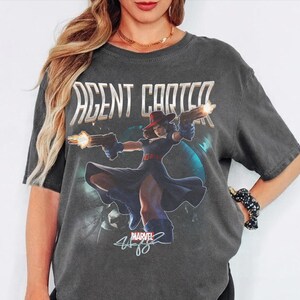 Pu&ograve; includere: T-shirt grigio scuro con la scritta "AGENT CARTER" sopra una grafica di una donna con cappello rosso e vestito blu che tiene due pistole. Il logo Marvel e una firma sono sotto.