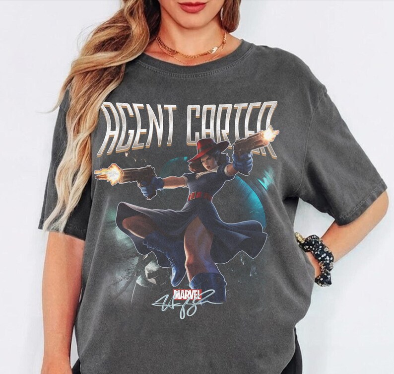 Pu&ograve; includere: T-shirt grigio scuro con la scritta "AGENT CARTER" sopra una grafica di una donna con cappello rosso e vestito blu che tiene due pistole. Il logo Marvel e una firma sono sotto.