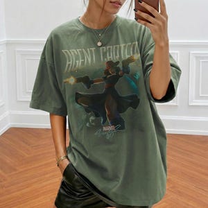 Pu&ograve; includere: T-shirt verde oliva con grafica di Agent Carter. La maglietta presenta un'illustrazione stilizzata di Agent Carter che tiene due pistole, con la scritta "AGENT CARTER" sopra l'immagine e "MARVEL" sotto. La maglietta &egrave; oversize.