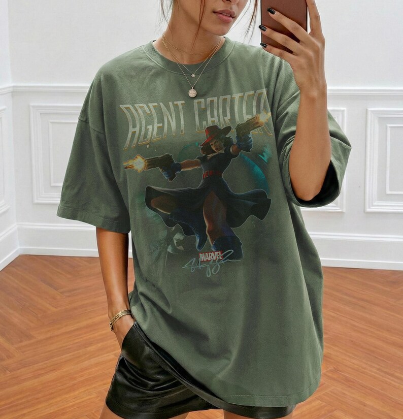 Pu&ograve; includere: T-shirt verde oliva con grafica di Agent Carter. La maglietta presenta un'illustrazione stilizzata di Agent Carter che tiene due pistole, con la scritta "AGENT CARTER" sopra l'immagine e "MARVEL" sotto. La maglietta &egrave; oversize.
