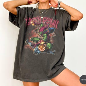 May include: Dark gray t-shirt featuring a Green Goblin graphic. The design includes the text "GREEN GOBLIN" in a distressed, pink font, and a detailed illustration of the Green Goblin character. The shirt has a vintage, worn look.