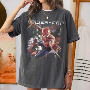 May include: A charcoal gray short-sleeve t-shirt featuring a graphic print of the character Spider-Man in a dynamic pose. The text "SPIDER-MAN" is printed in white capital letters above the character. The shirt has a crew neckline and a relaxed fit.