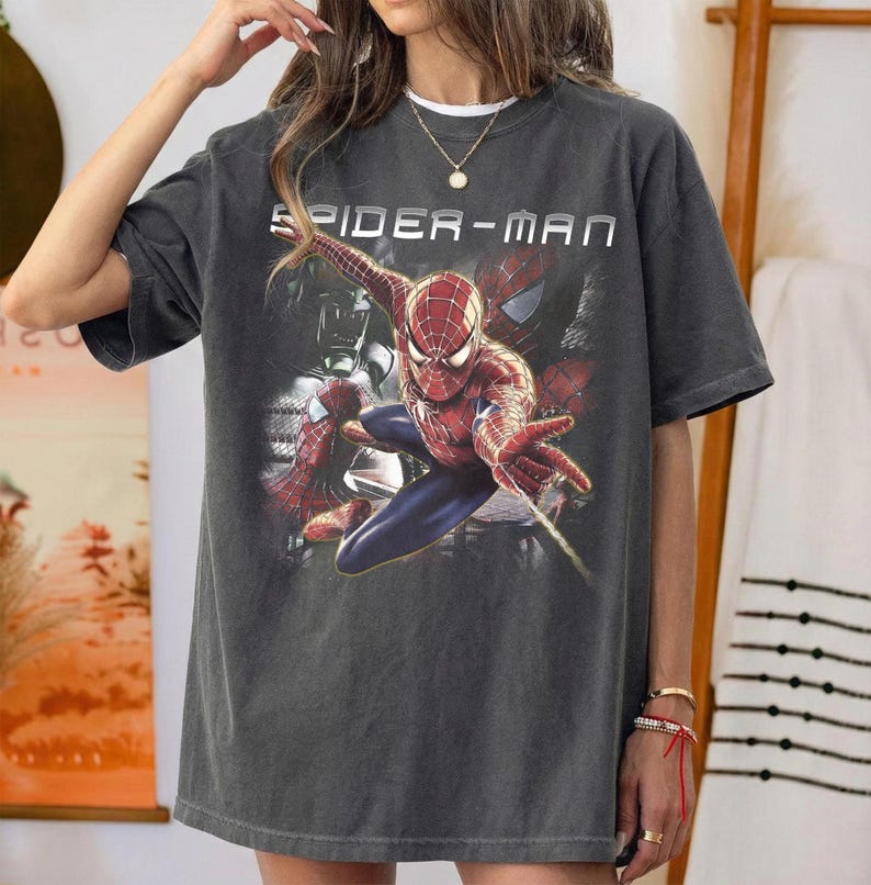 May include: A charcoal gray short-sleeve t-shirt featuring a graphic print of the character Spider-Man in a dynamic pose. The text "SPIDER-MAN" is printed in white capital letters above the character. The shirt has a crew neckline and a relaxed fit.