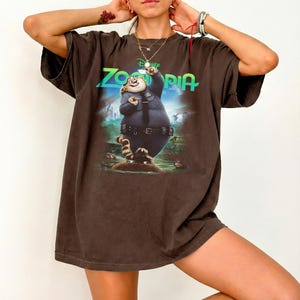 K&ouml;nnte beinhalten: Ein dunkelbraunes Oversize-T-Shirt mit einem Grafikdruck der Figur Officer Clawhauser aus dem Film Zoomania. Das Design zeigt das leuchtend gr&uuml;ne Zoomania-Logo &uuml;ber der Figur, die eine blaue Polizeiuniform tr&auml;gt.