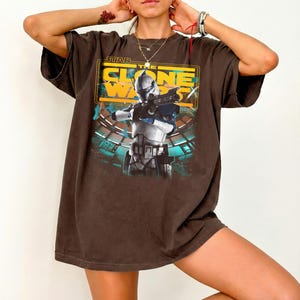 May include: A dark brown short-sleeve t-shirt featuring a graphic print of a Star Wars The Clone Wars character in white and blue armor holding a blaster. The design includes yellow text and a teal background with a circular structure.