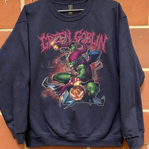 May include: Navy blue sweatshirt featuring a graphic of the Green Goblin character. The design includes the text "GREEN GOBLIN" in a stylized font, along with an illustration of the character in action. The sweatshirt is a crew neck style.
