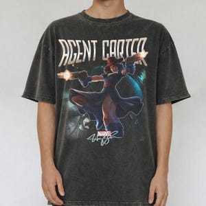 Pu&ograve; includere: T-shirt grigio scuro di Agent Carter con una grafica Marvel del personaggio con le pistole. La maglietta presenta la scritta "AGENT CARTER" in oro e una firma. La maglietta ha un aspetto vintage.