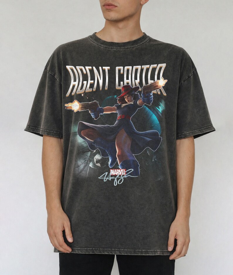 Pu&ograve; includere: T-shirt grigio scuro di Agent Carter con una grafica Marvel del personaggio con le pistole. La maglietta presenta la scritta "AGENT CARTER" in oro e una firma. La maglietta ha un aspetto vintage.
