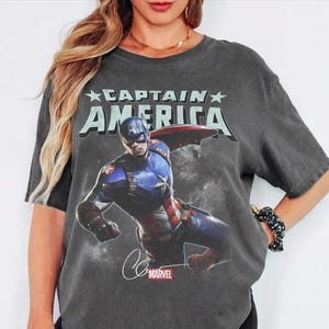 Puede incluir: Camiseta gris oscuro con un gr&aacute;fico del Capit&aacute;n Am&eacute;rica. El texto "CAPTAIN AMERICA" est&aacute; impreso encima de la imagen, con el logotipo de Marvel y la firma debajo. La camiseta tiene un aspecto vintage y desgastado.