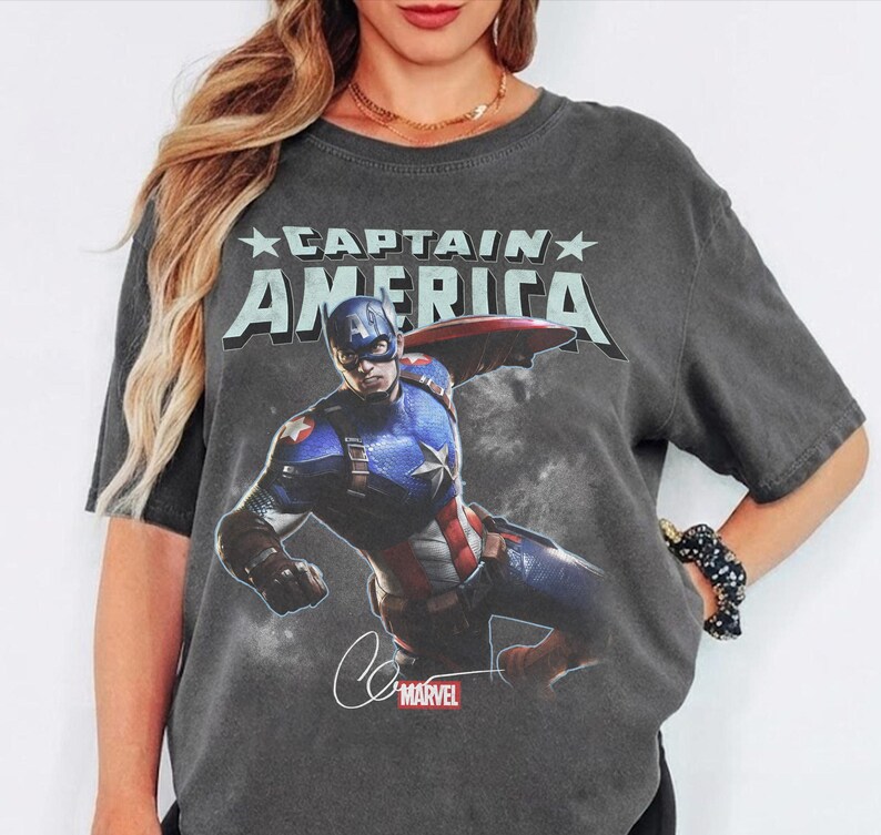 Puede incluir: Camiseta gris oscuro con un gr&aacute;fico del Capit&aacute;n Am&eacute;rica. El texto "CAPTAIN AMERICA" est&aacute; impreso encima de la imagen, con el logotipo de Marvel y la firma debajo. La camiseta tiene un aspecto vintage y desgastado.