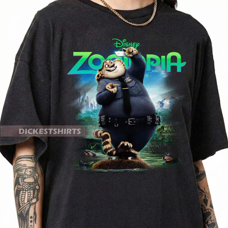 K&ouml;nnte beinhalten: Ein schwarzes T-Shirt mit einem Grafikdruck der Figur Chief Bogo aus dem Film Zoomania. Die Figur tr&auml;gt eine dunkelblaue Polizeiuniform, h&auml;lt ein kleines wei&szlig;es Ger&auml;t und steht &uuml;ber einem braunen, pelzigen Wesen.