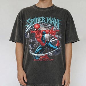 May include: Dark gray t-shirt featuring a graphic of Spider-Man in action, with the words "SPIDER MAN" in blue and "BRAND NEW DAY" below. The superhero is depicted in his classic red and blue suit, set against a cityscape background.