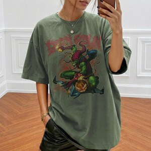 May include: Olive green t-shirt featuring a graphic of a green goblin-like character with a pumpkin and the text "Dead Run" in red. The design is centered on the front of the shirt. The shirt has short sleeves and a relaxed fit.
