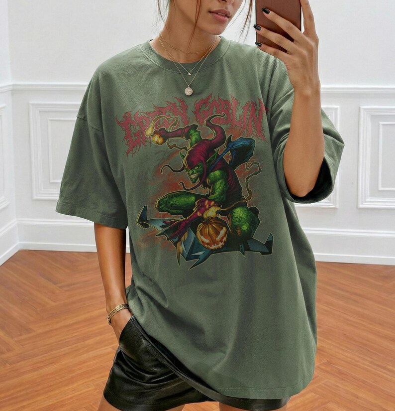 May include: Olive green t-shirt featuring a graphic of a green goblin-like character with a pumpkin and the text "Dead Run" in red. The design is centered on the front of the shirt. The shirt has short sleeves and a relaxed fit.