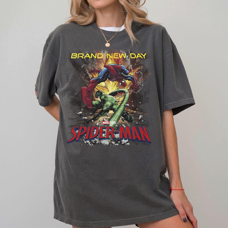 May include: A charcoal gray short-sleeve t-shirt featuring a graphic print of Spider-Man and a green creature in a city scene. The text "BRAND NEW DAY" appears in yellow above the characters, and "SPIDER-MAN" is printed in large red letters below.