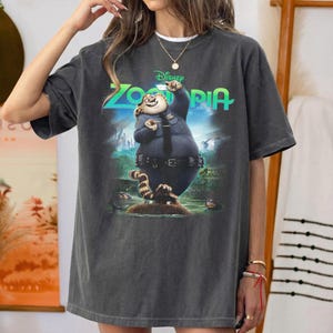 K&ouml;nnte beinhalten: Ein anthrazitfarbenes Kurzarm-T-Shirt mit einem Grafikdruck der Figur Officer Clawhauser aus dem Film Zoomania. Das Design zeigt das gr&uuml;ne Zoomania-Logo &uuml;ber der Figur, die eine blaue Polizeiuniform tr&auml;gt.